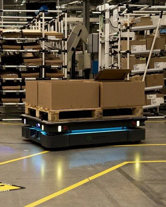 Fabrika otomasyonu kapsamında sarı şerit çizgili üretim alanında koli yüklü paleti taşıyan otonom mobil robot, arkada depolama rafları